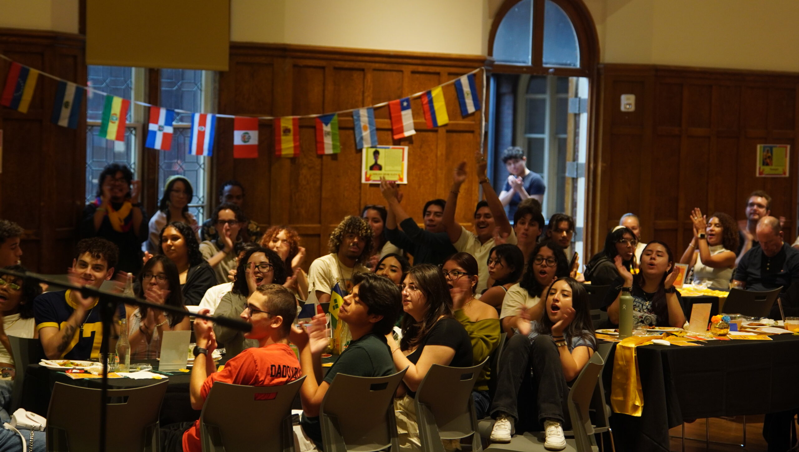 Large group of students seated in chairs looking ahead at the performers. Some students have their hands in the air cheering and clapping.