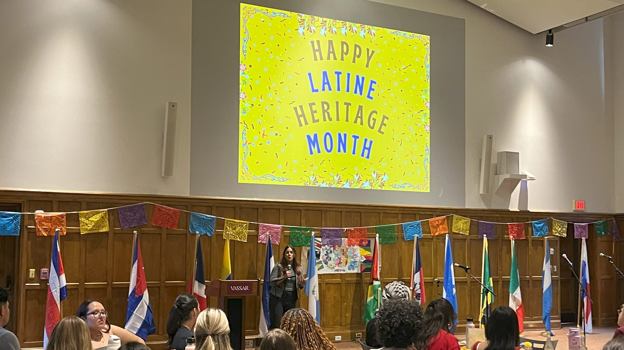 Community Guest standing at the from of the room speaking in the mic with the words "Happy Latine Heritage Month" in the background.