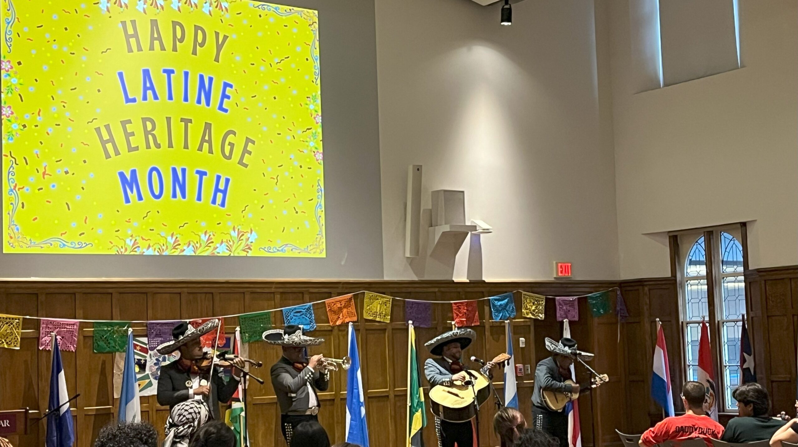 Group of four mariachi band members standing side by side in front of the audience as they perform each with a different instrument in their hands as each member is wearing a sombero on their head. 