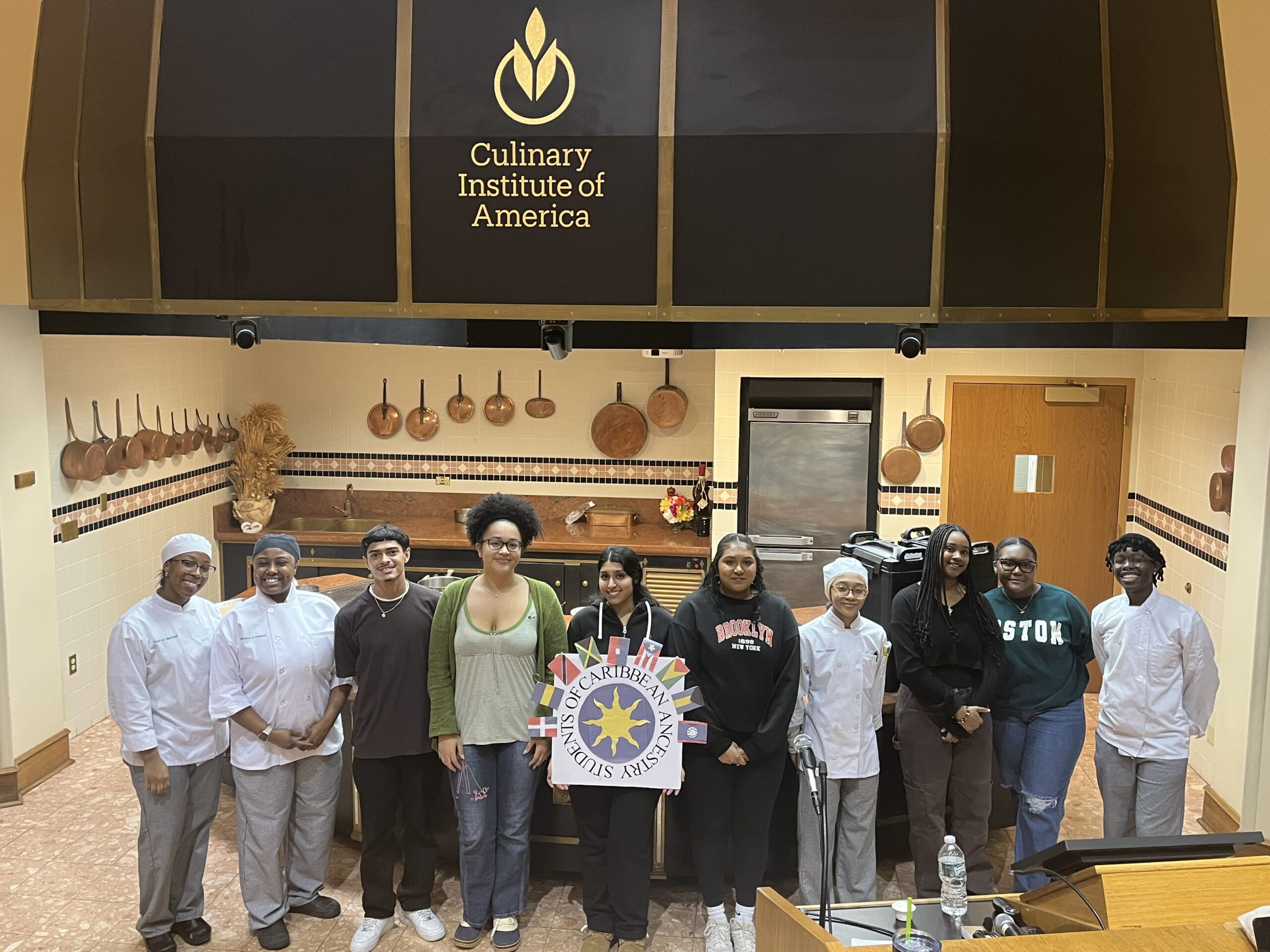 Ten student leaders standing side by side in a line. The student in the center is holding up a square logo with Caribbean flags around the words "Students of Caribbean Ancestry." The students are standing under a sign that reads "Culinary Institute of America."