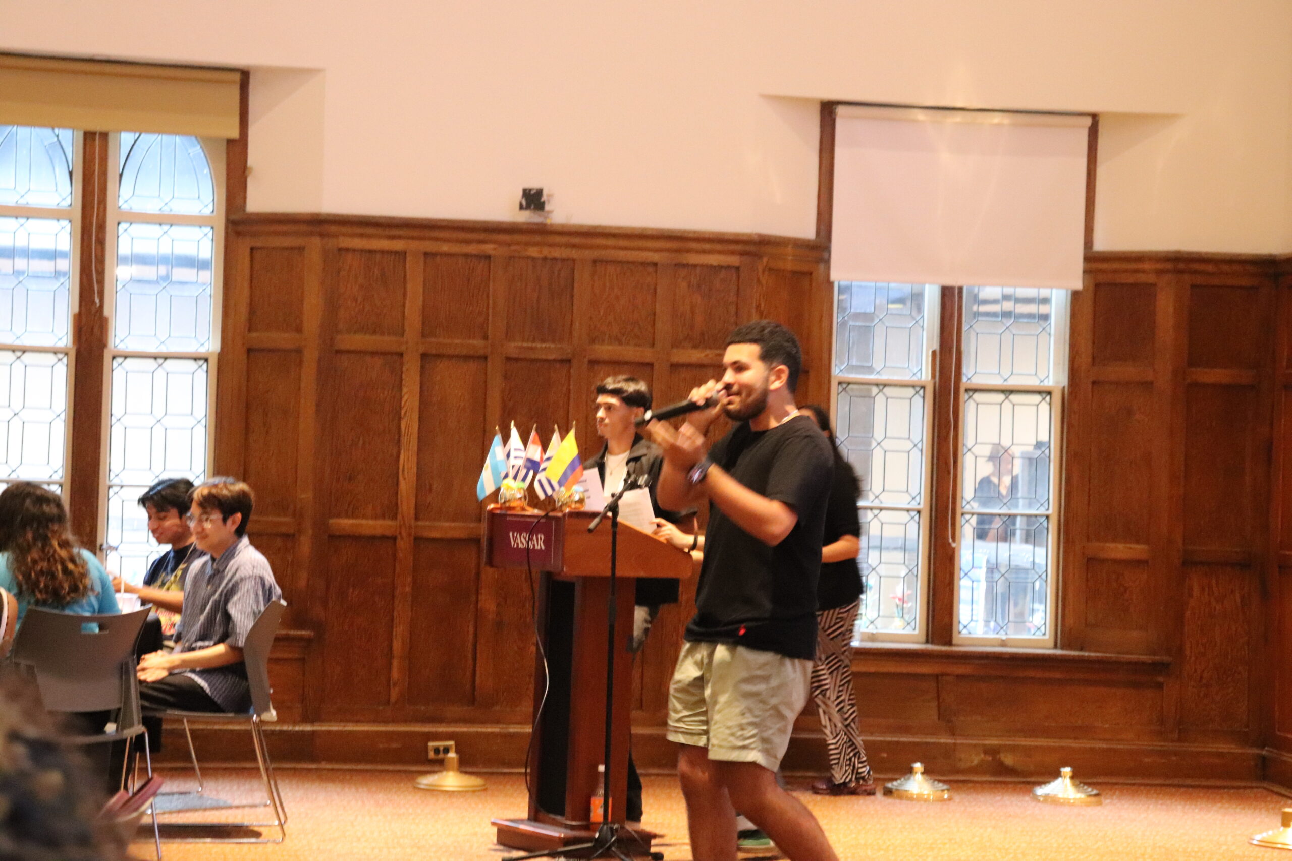 Student speaking into the mic as his left arm up and his right hand is holding the mic. Students are seated to the left of the image and two students are pictured behind the student speaking.