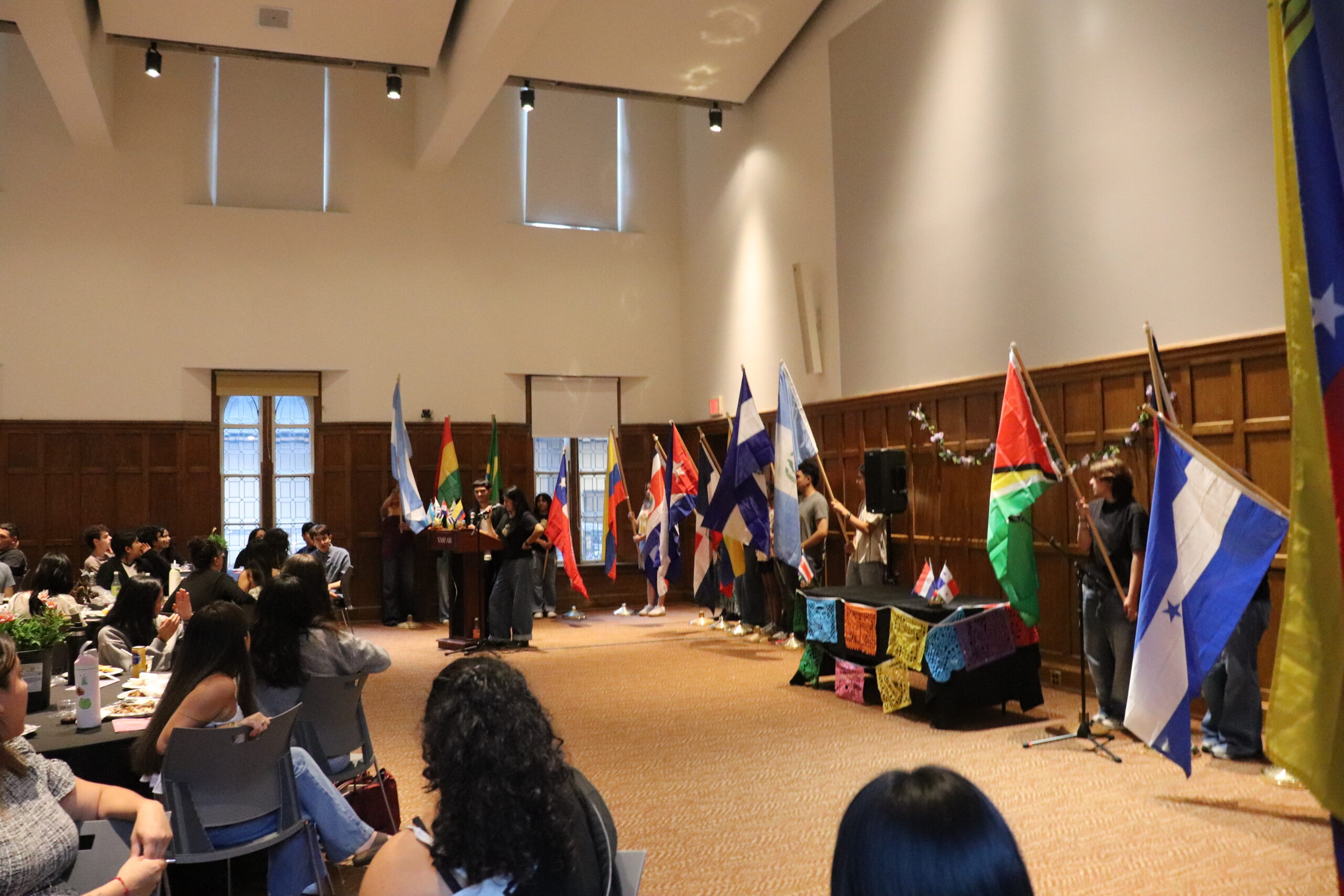 Room filled with students lined up with flags in their hands as two students stand behind the podium speaking into the mic as the audience looks on.