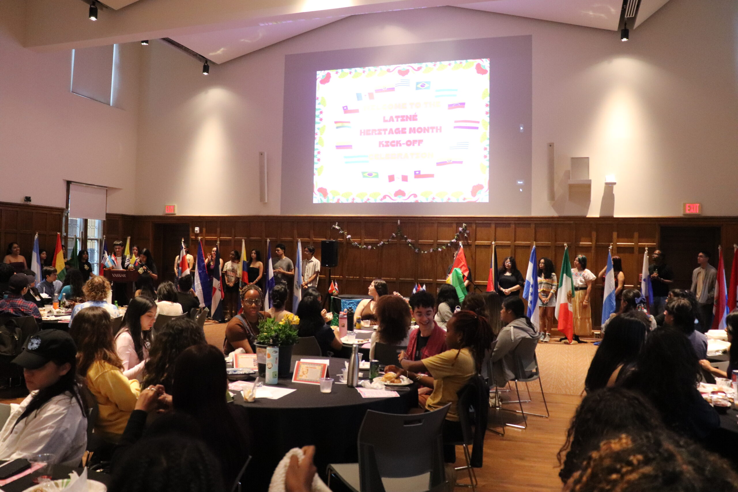 Room filled with students seated at round tables and students standing next to the flag poles as an image is displayed on the large screen in the middle of the image highlighting "Latine Heritage Month Kick-off."