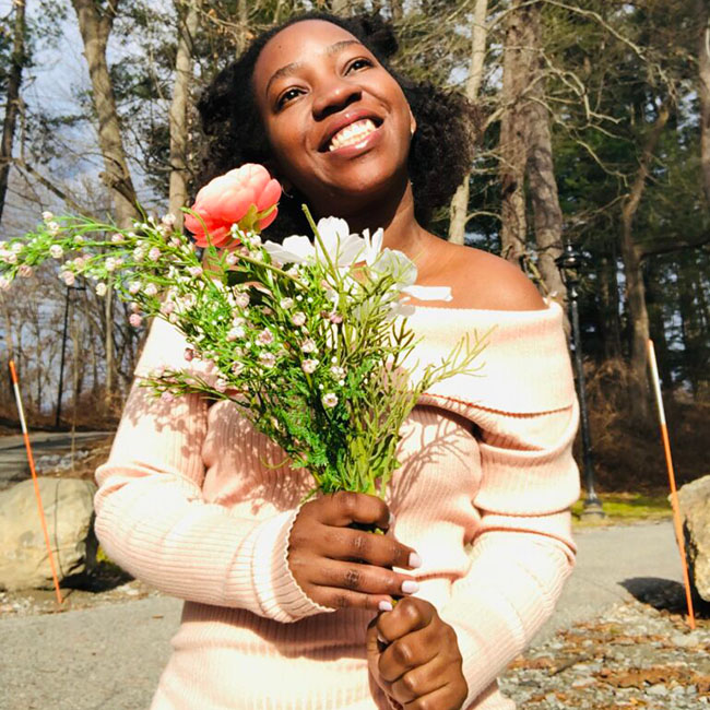 Photo of Breille smiling, looking up while holding a bouquet of flowers with both hands as she stands in front of a group of trees and rocks off to each side.  