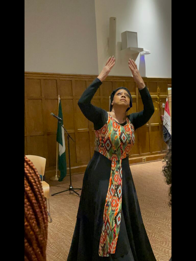 A woman in African attire standing in a pose with her hands over her head.