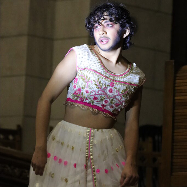 A photo of Kiran posing with they're head to the left. Kiran is wearing a white two piece outfit with pink and gold details. 
