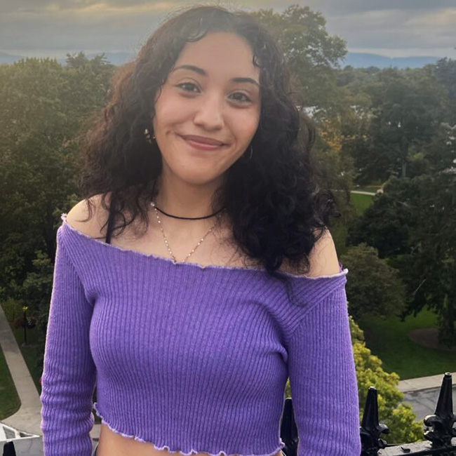 Photo of Sofia standing in front of a group of green filled trees as she wears a purple long-sleeved shoulder crop top as she smiles into the camera. 
