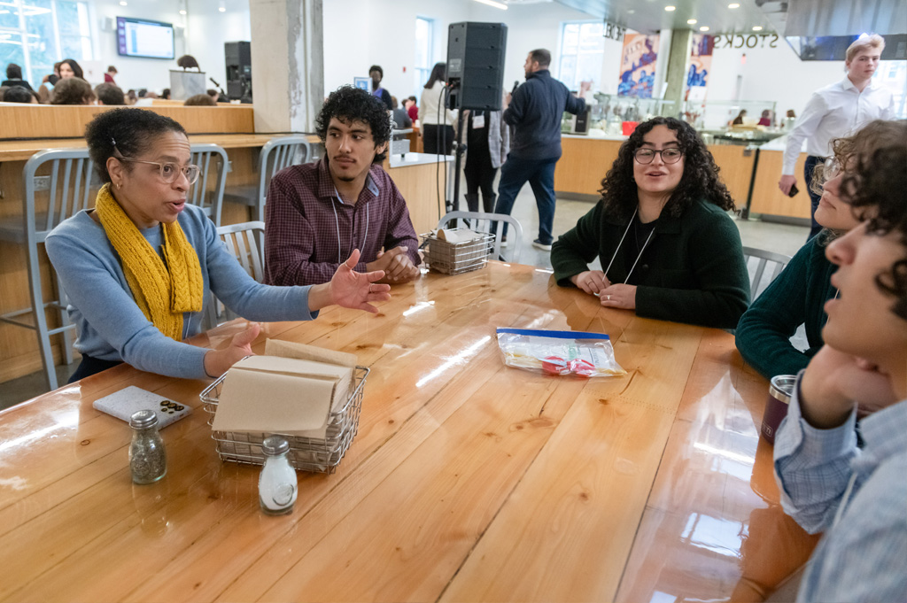 Students and a staff member engaged in a group discussion at a table in a campus common area.