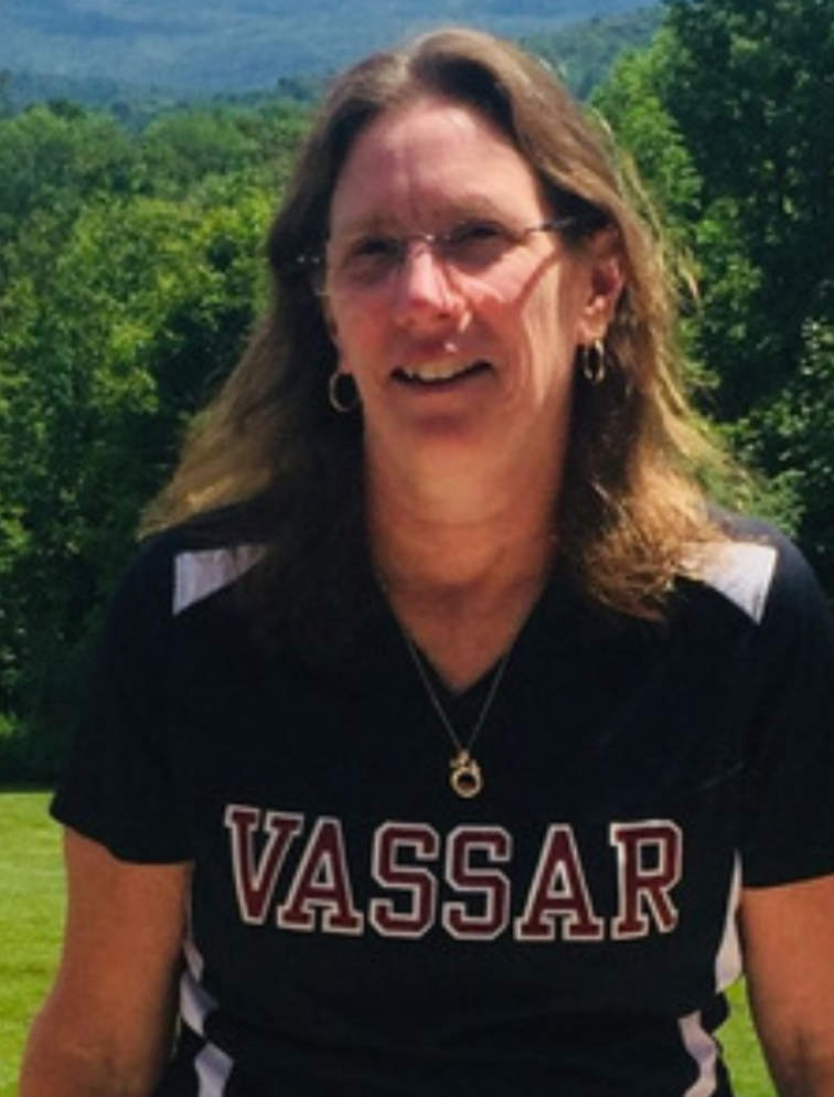 A photo portrait of Andrea Pesavento, a person with glasses, long blond hair, and a black T-shirt that reads "Vassar". They are standing outside and smiling at the camera.