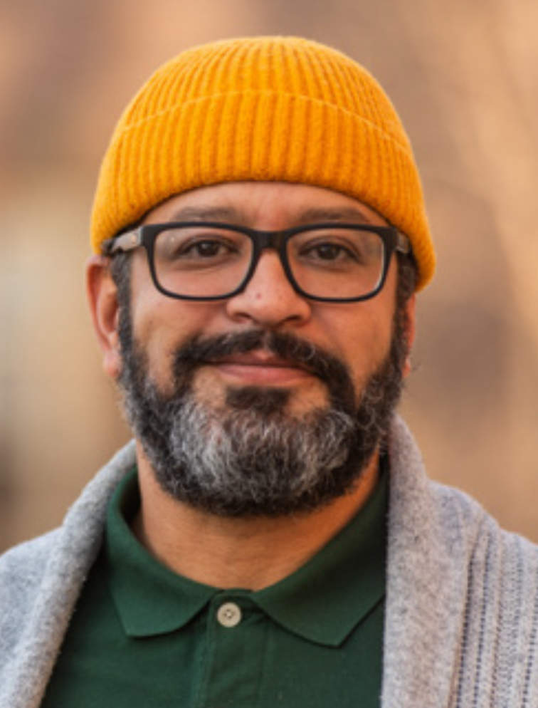 A photo portrait of Luis Jiminez Inoa, a person with an orange hat, glasses, a gray beard, and a green shirt. They are smiling at the camera.