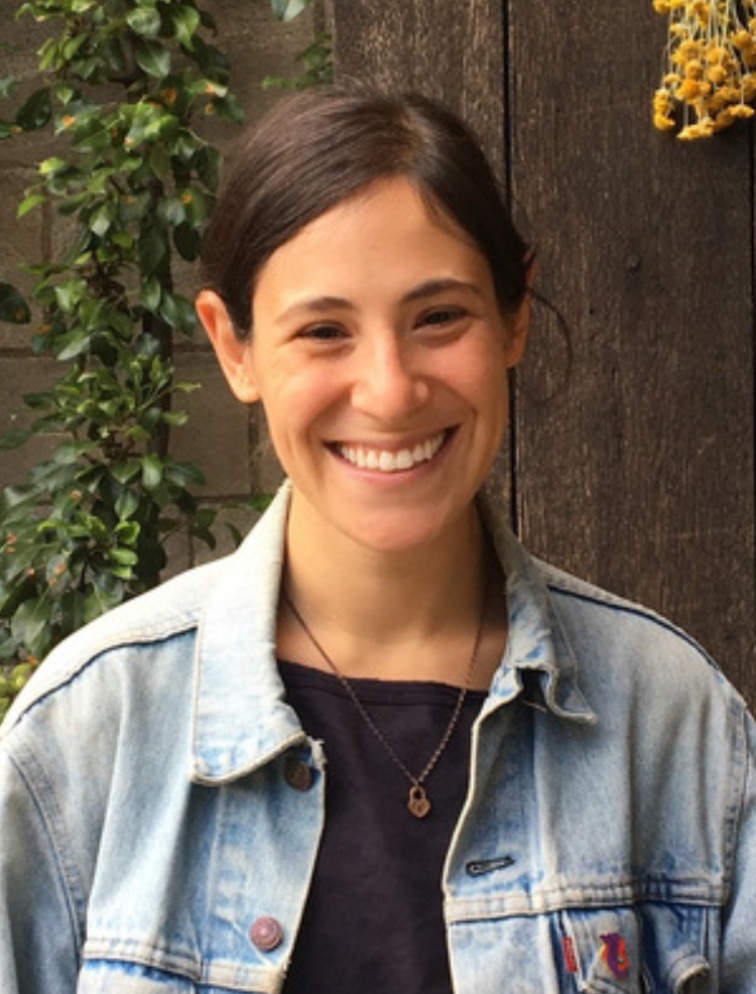 A photo portrait of Rachel Gellert, a person with long brown hair and a blue denim shirt. They are smiling at the camera.
