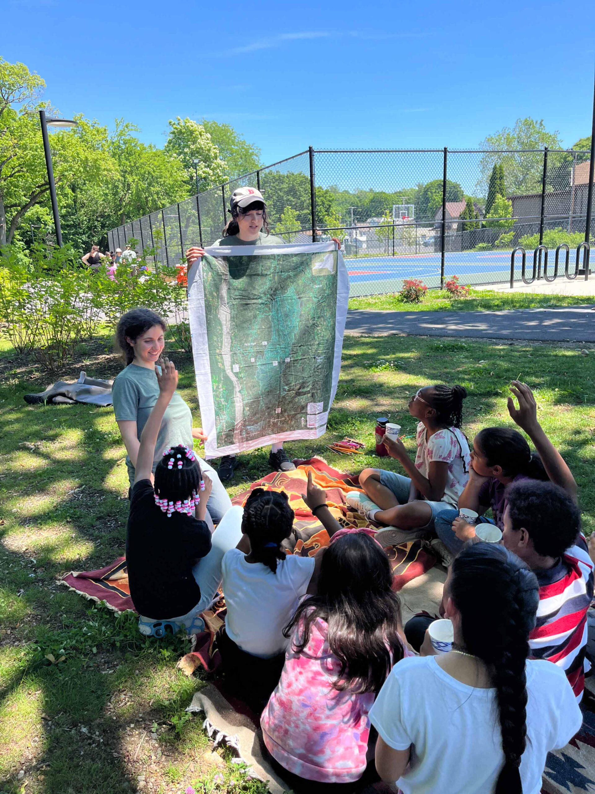 A group of children sitting on a blanket outdoors on a sunny raise their hands while an instructor holds up a large map.