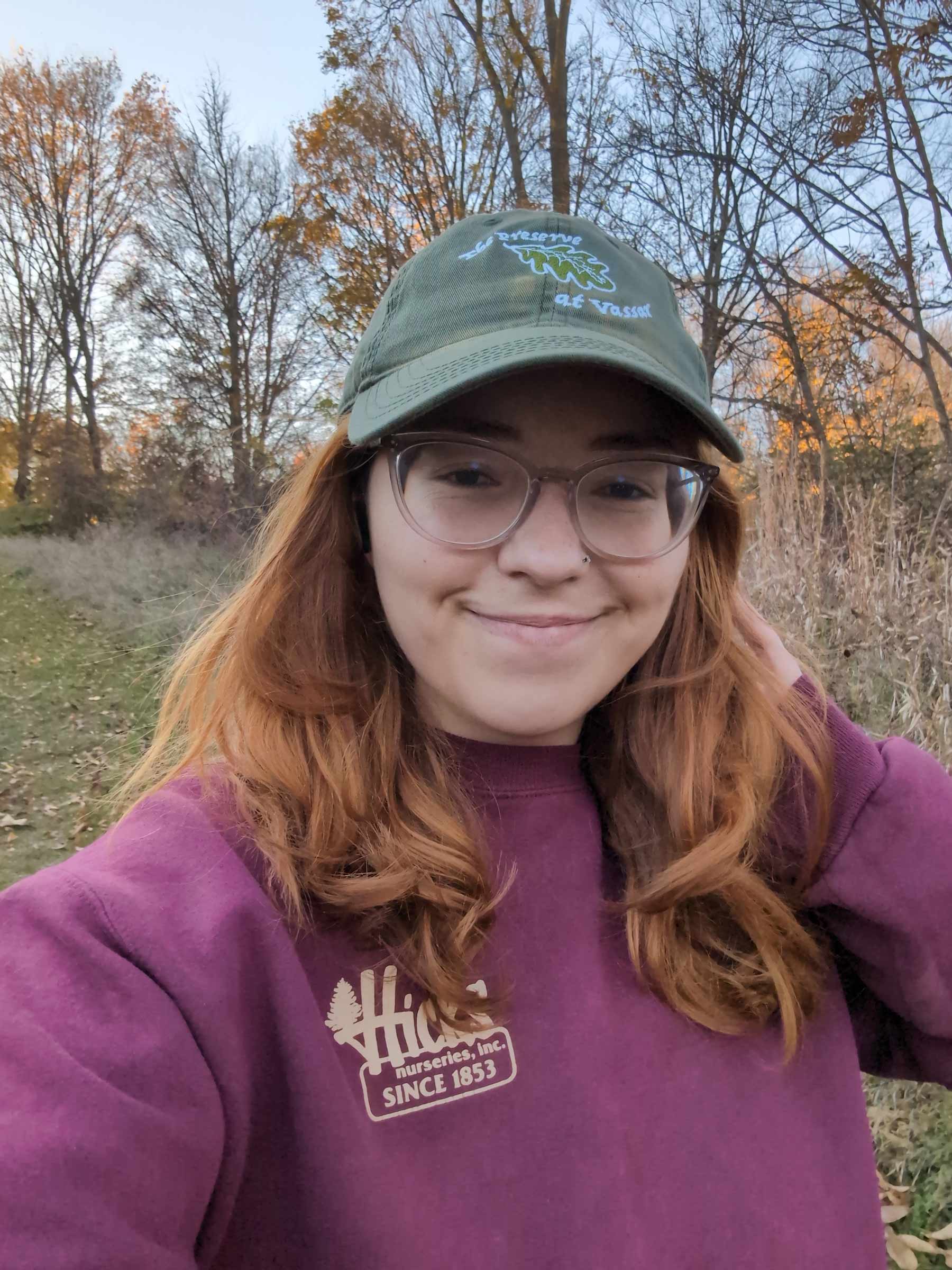 An individual with long reddish-brown hair wearing glasses and an olive green baseball cap smiling outdoors. They are wearing a maroon sweatshirt with a logo. The background shows a field and bare trees under a clear sky.