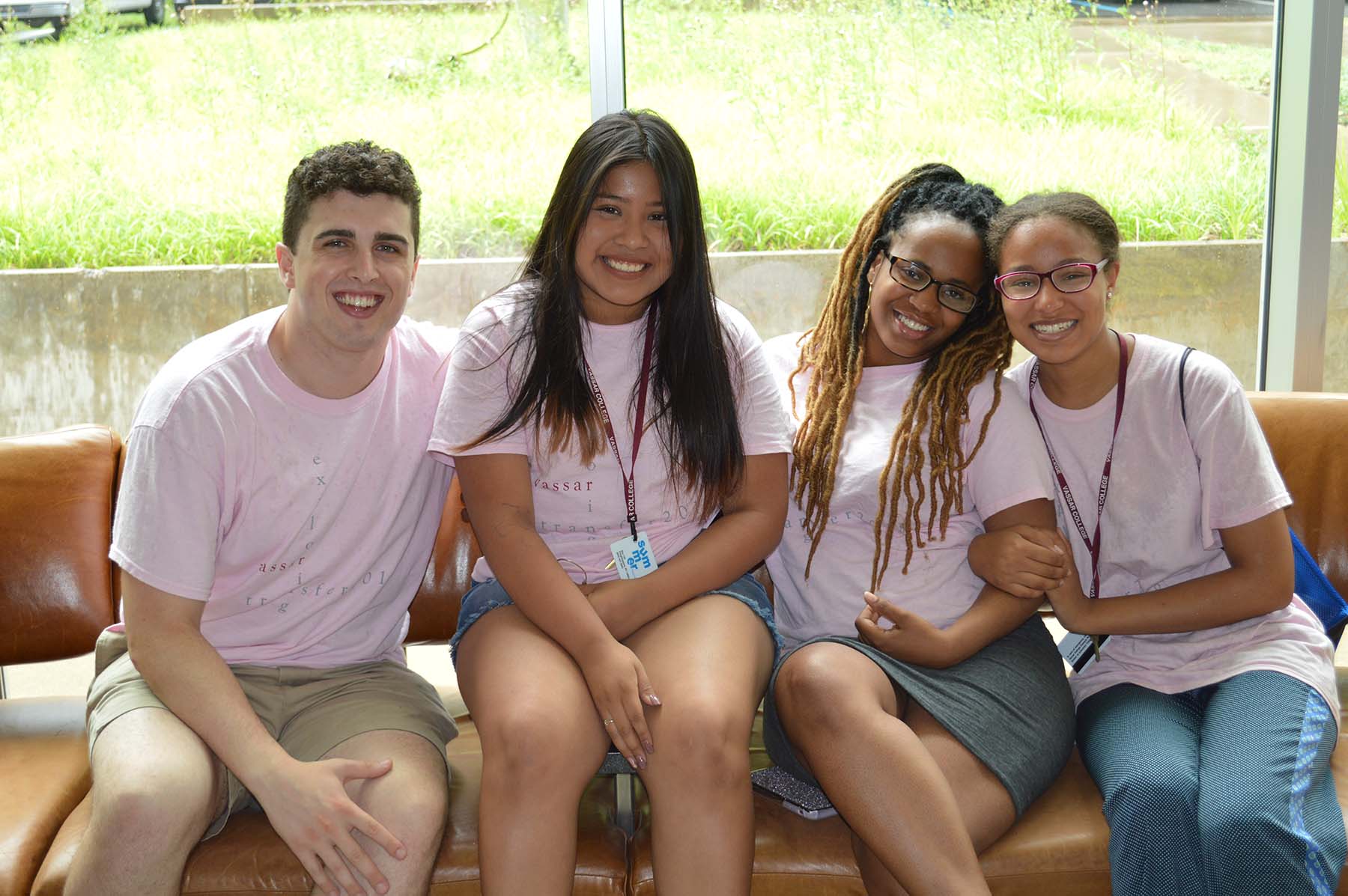 Four people wearing pink t-shirts sitting on a beige colored couch in front of a wall of windows.