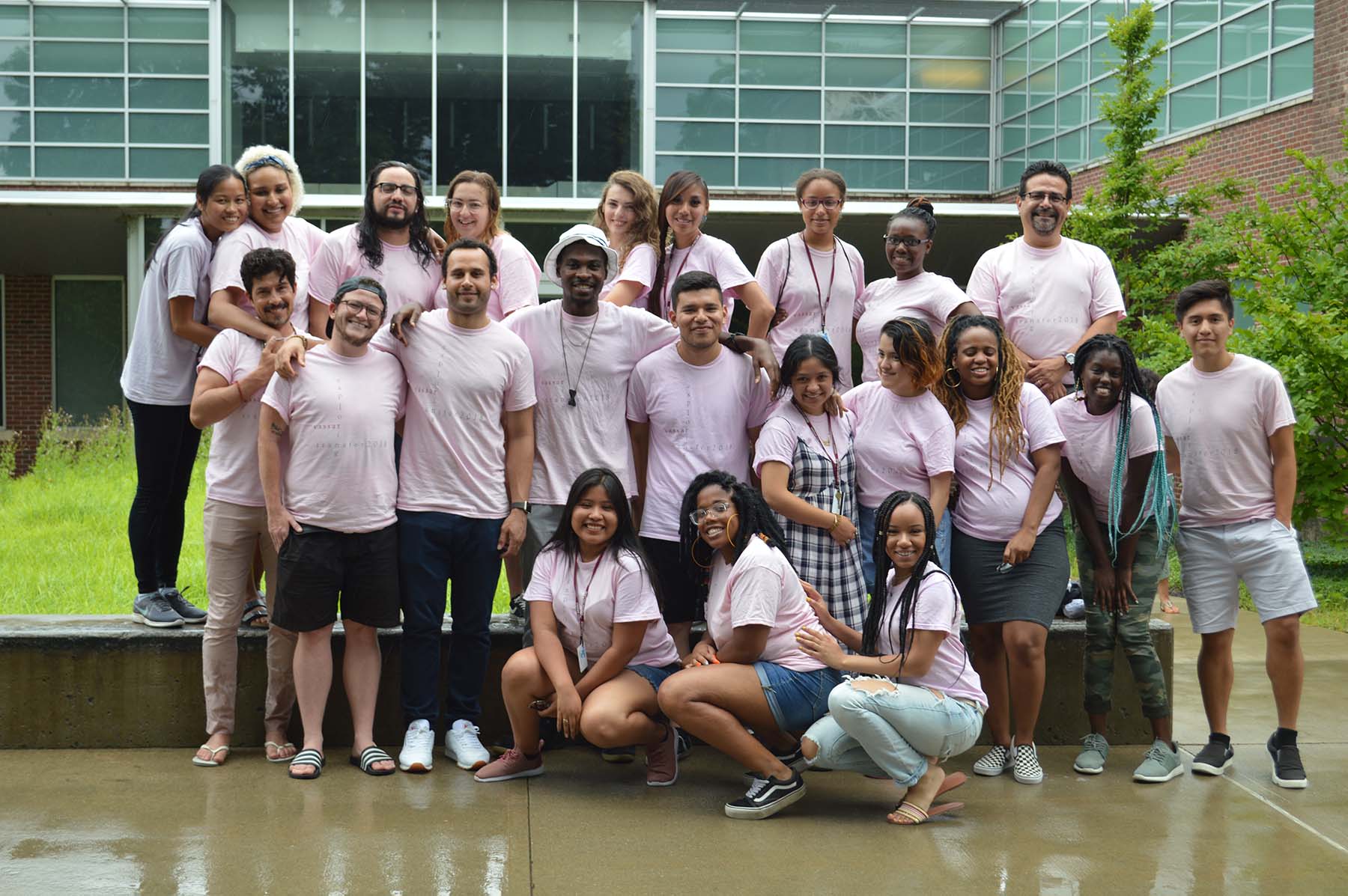 Large group of people all wearing light pink t-shirts in front of a building with large windows.