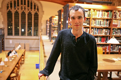 Alexander "Sasha" Steinberg ’09 with short dark hair wearing a black sweater, standing in a warmly lit library with tall bookshelves and a large arched window in the background.