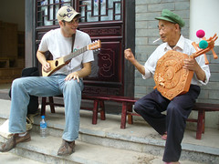 Two men sitting on steps, including Jonathan Kaiman ’09, playing string instruments, smiling at each other. One wears a baseball cap, t-shirt, and jeans while playing a guitar-like instrument; the other wears a green cap, white shirt, and dark pants while playing a traditional round instrument decorated with carvings and colorful tassels.