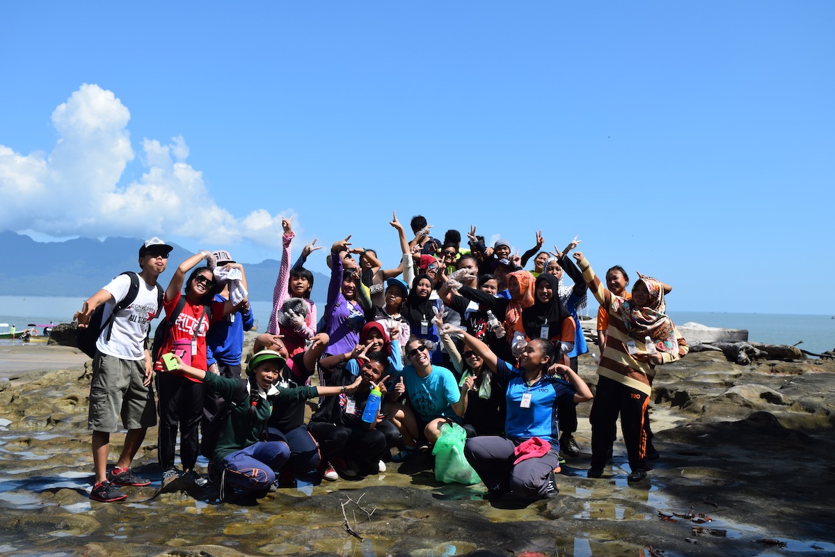 Eleni Macrakis’ 14 with a large group of smiling people at Bako National Park, Malaysia, on rocky terrain by the ocean under a bright blue sky, with mountains visible in the distance. Many are making playful gestures and peace signs, wearing casual outdoor clothing and hats.