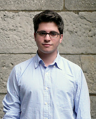 Michael Frenkel ’09 with short dark hair and glasses wearing a light blue button-up shirt, standing against a textured stone wall.