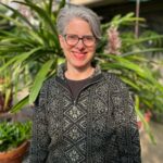 Jessica-Greenstein smiling with short gray hair and glasses, wearing a patterned black and gray sweater, standing in front of lush green plants.