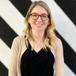 Kerry Zuccaro smiling with long blonde hair and glasses, wearing a black top and beige cardigan, standing in front of a black and white striped wall.