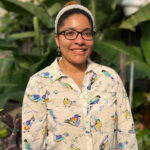 Yamelli-Hernandez smiling with glasses and a pearl headband, wearing a cream-colored shirt decorated with colorful bird illustrations, standing in front of large green plants.
