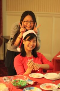 Two women at a table decorating cookies with one behind the other making "bunny ears" on her head.