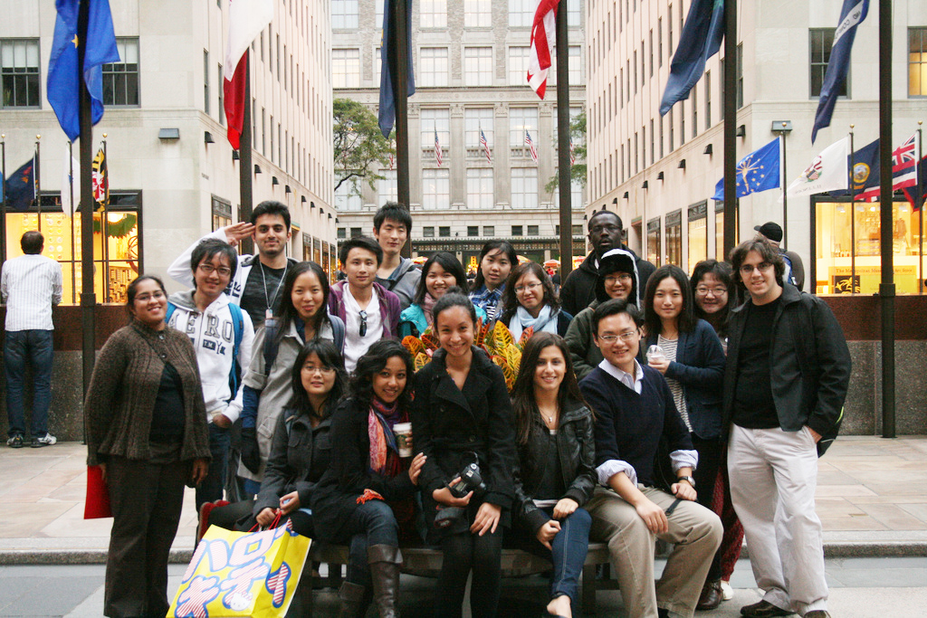 People posing in front of large buildings with flags of many countries.