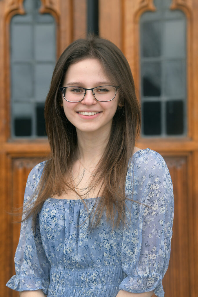 Jackie Rosenblum with long brown hair and glasses wearing a light blue floral dress, smiling in front of wooden doors with glass panels.