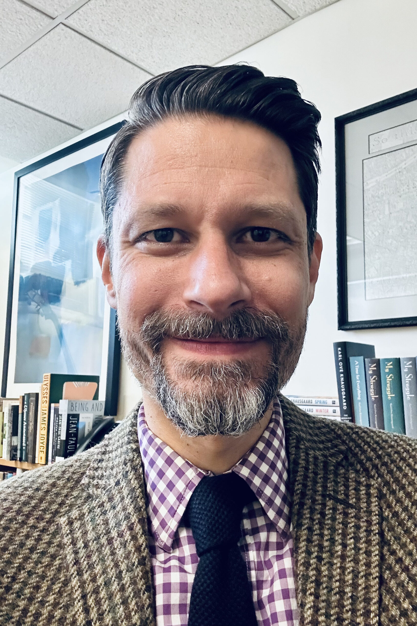 Matthew Schultz smiling in an office setting, wearing a brown tweed jacket, a purple and white gingham dress shirt, and a dark knit tie.