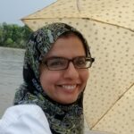 Portrait of a person outdoors by the water, smiling beneath a patterned umbrella, wearing glasses and a headscarf.