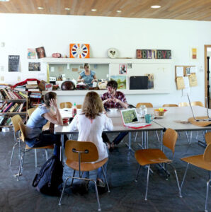Three people sitting around a large table in a brightly lit common room with a wood-paneled ceiling, talking and working on laptops, while another person cooks in the open kitchen area in the background.