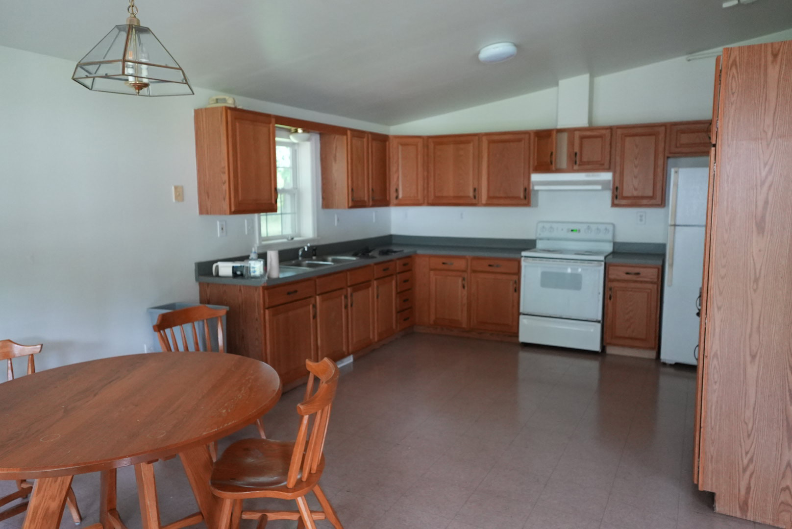 Two walls lined with wood kitchen cabinets, sink, white oven, white refrigerator, and a round wood table and chairs.