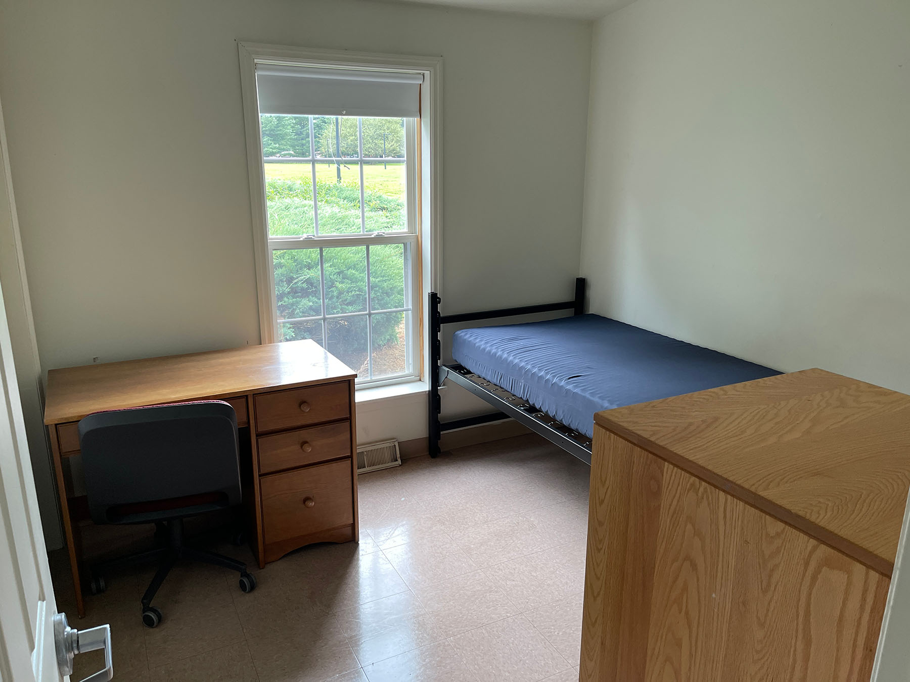 Room with a small wood desk, black rolling chair, bed frame and mattress and wood dresser.
