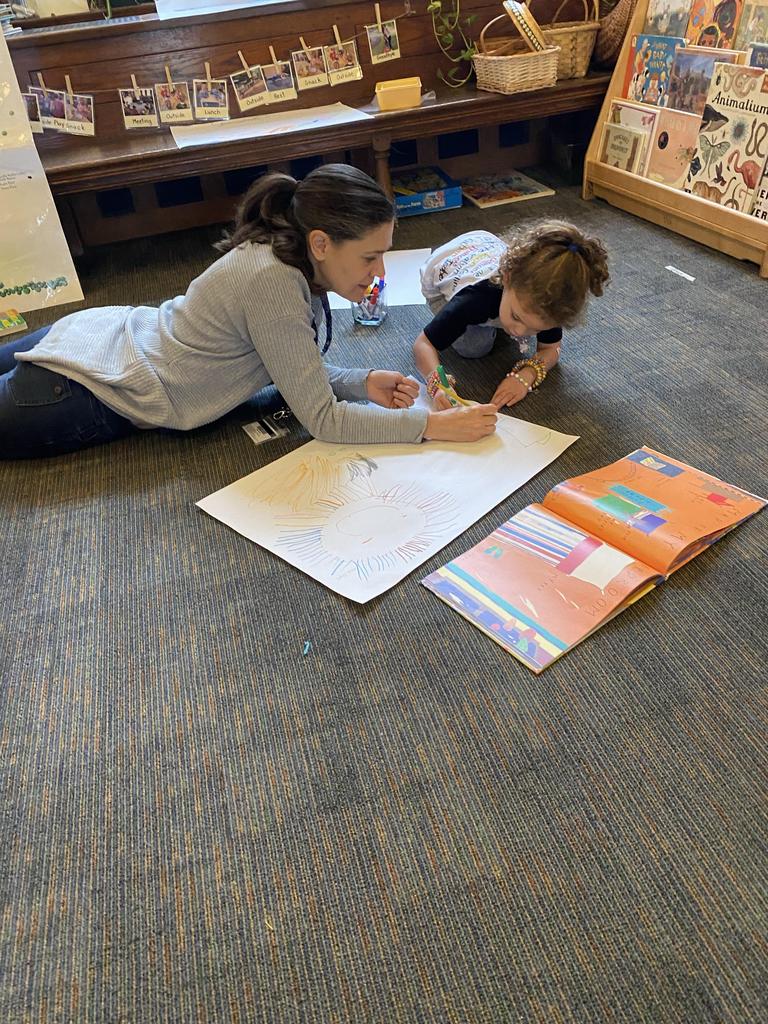 An adult and a small child sit on the floor together, looking at a large sheet of paper. The child is speaking, and the adult is writing it down.