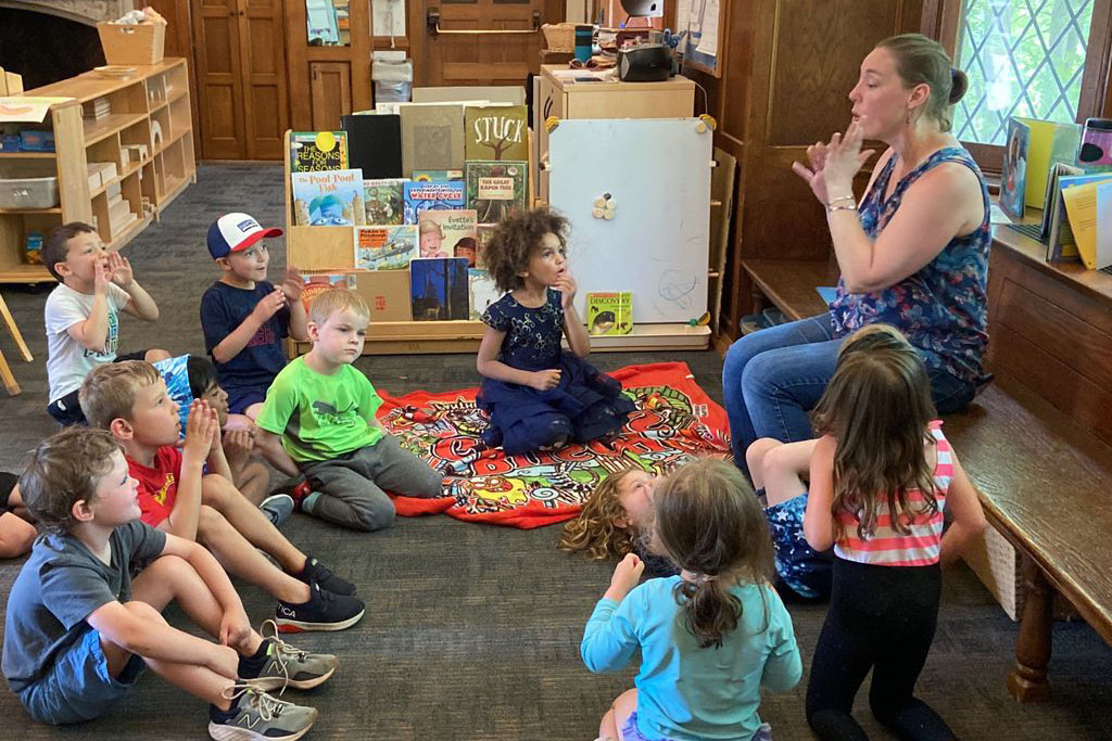 Adult using hand gestures while small children sit on a colorful rug and listen in a classroom with books displayed behind them.