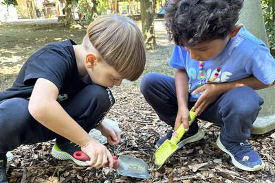 Two small children crouched outdoors using hand trowels to dig in wood-chip covered ground.