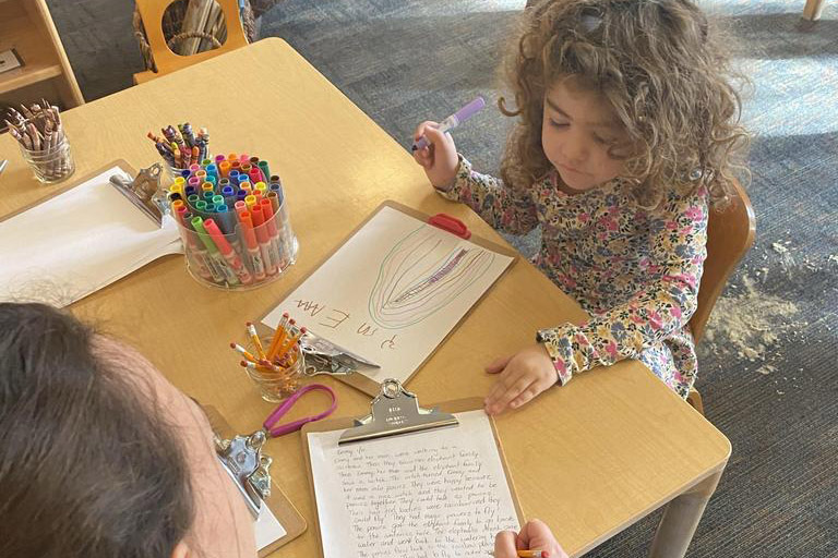 Small child drawing with markers at a table while an adult writes on a clipboard nearby.