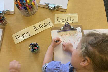 Small child writing “Happy Birthday Daddy” on paper at a table with markers and a matching nameplate.