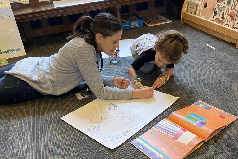 Adult lying on the floor drawing with a small child who is coloring on a large piece of paper, an open book nearby in a classroom.