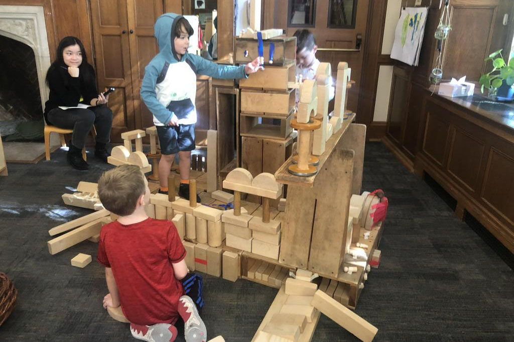 Small children assemble a large wooden block structure on a classroom floor while a person watches from a chair in the background.