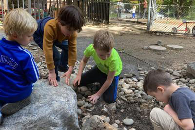 Four small children crouch and kneel around a rocky patch outdoors, examining and moving stones near a large boulder.