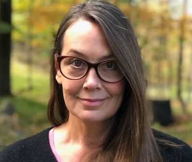 A headshot of Carol Garboden Murray, a person with long brown hair and glasses, standing in a wooded area, looking at the viewer.