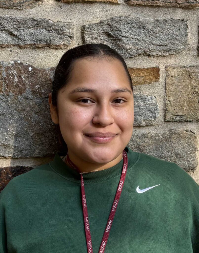 A close-up portrait of an individual wearing a green sweatshirt features a prominent white logo on the chest. The background consists of textured stone walls in various shades of gray and brown, providing a natural contrast. A lanyard with a maroon ribbon bearing the word "VASSAR" hangs around the neck, accompanied by a small purple accessory. The lighting is soft and natural, highlighting the facial features and creating gentle shadows against the backdrop.