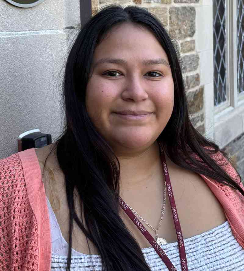 A person, Kimberly Reyes, is positioned in the foreground, facing the viewer with a slight smile. Long, dark hair flows over one shoulder, contrasting with a light, patterned top that features horizontal stripes. They wear a coral cardigan and a lanyard with the word "Vassar" printed on it. The background includes a textured stone wall and windows that add architectural detail, with natural lighting enhancing the scene's clarity and warmth.