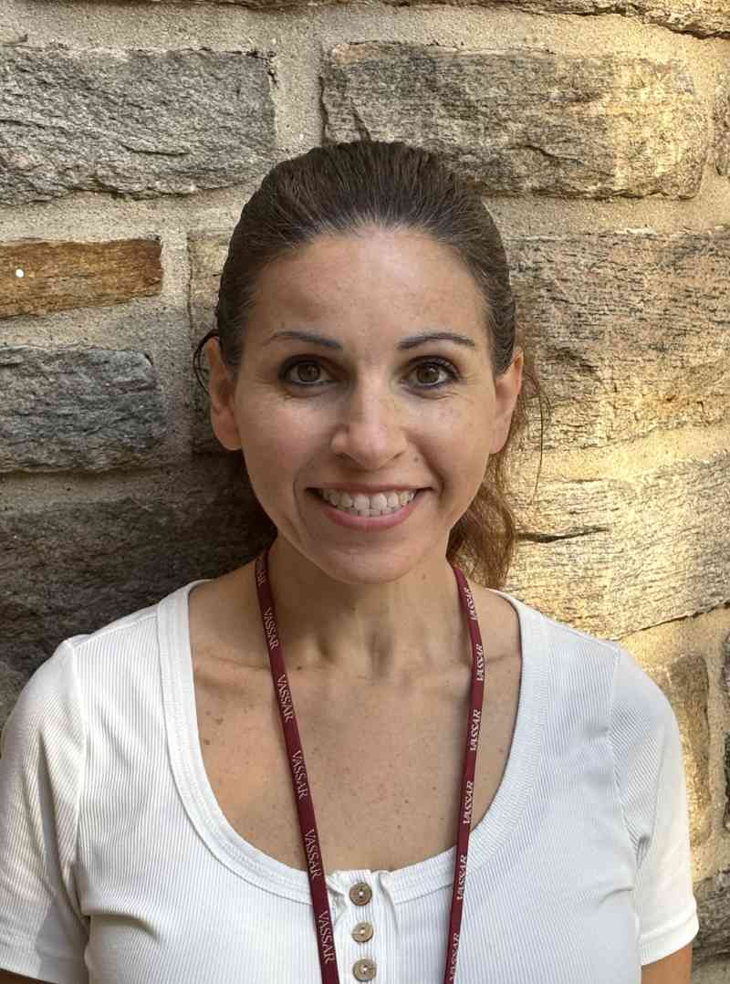 A person, Tara Nelgia, stands smiling in front of a textured stone wall, which features a mix of light and dark stones creating a natural pattern. They are wearing a white shirt with a subtle ribbed texture and a lanyard that has a dark maroon color and repeating text. Warm, diffused light from an off-camera source highlights their features and adds a gentle glow to the stones. The overall composition includes the figure centered against the rugged backdrop, creating a harmonious visual contrast.
