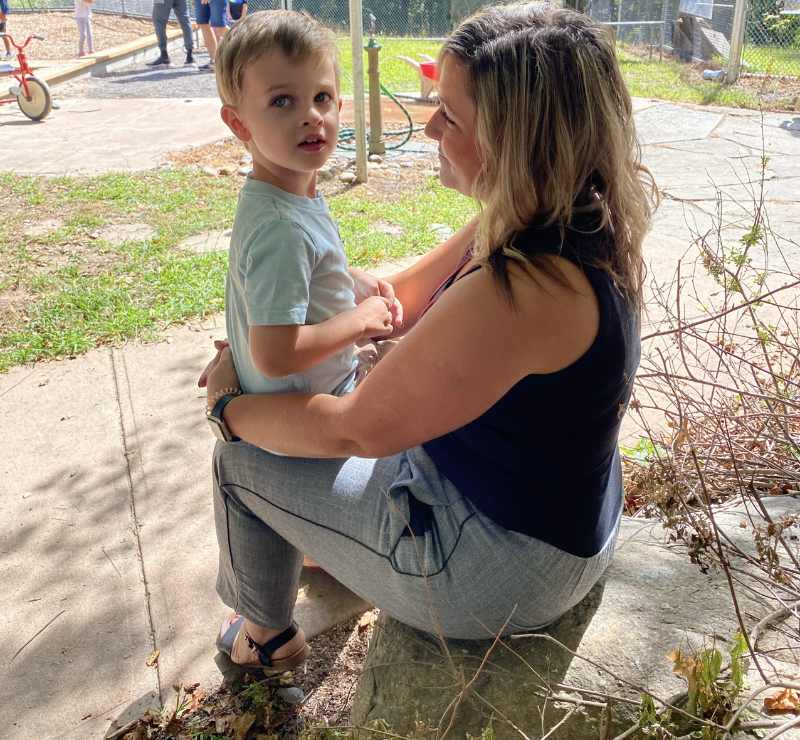 A figure sits on a large rock, holding a child facing them. The child, in a light blue shirt, appears inquisitive, with wide eyes directed slightly off-camera. The adult figure, wearing a dark tank top and light-colored pants, has long hair cascading over one shoulder and a gentle posture. In the background, a playground is visible, with children playing and sunlight filtering through lush green grass, creating a lively outdoor atmosphere.