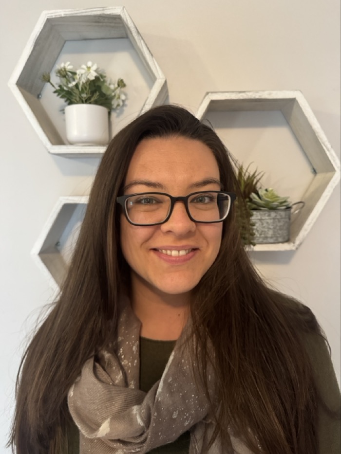The image features a person with long dark hair and glasses, smiling directly at the camera. They are dressed in a green top, paired with a textured scarf that has a light pattern. In the background, geometric hexagonal shelves display various plants and decor, adding a natural element to the scene. The lighting is soft and even, creating a warm atmosphere accentuated by the neutral wall.