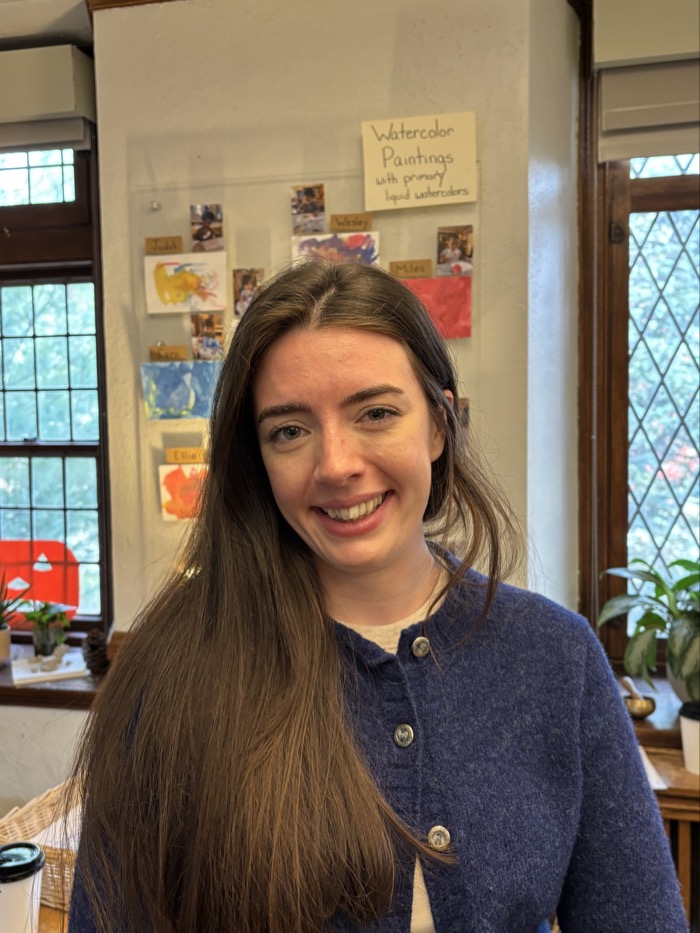 A person with long, dark hair stands smiling in a warmly lit room. They are wearing a navy blue cardigan with buttons, and the background features several colorful watercolor paintings pinned to a wall. Natural light filters through the nearby window, illuminating a potted plant on a table. A sign above the paintings indicates that they are created using primarily liquid watercolors.