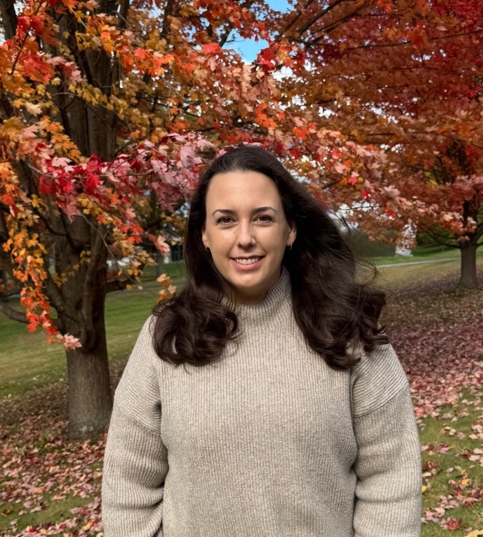 A figure stands in a grassy area, framed by vibrant autumn foliage displaying shades of red, orange, and yellow. The figure is positioned centrally, wearing a light gray, chunky knit sweater. Gentle sunlight illuminates the scene, casting soft shadows on the ground covered with fallen leaves. In the background, a blurred horizon hints at rolling hills, adding depth to the composition.